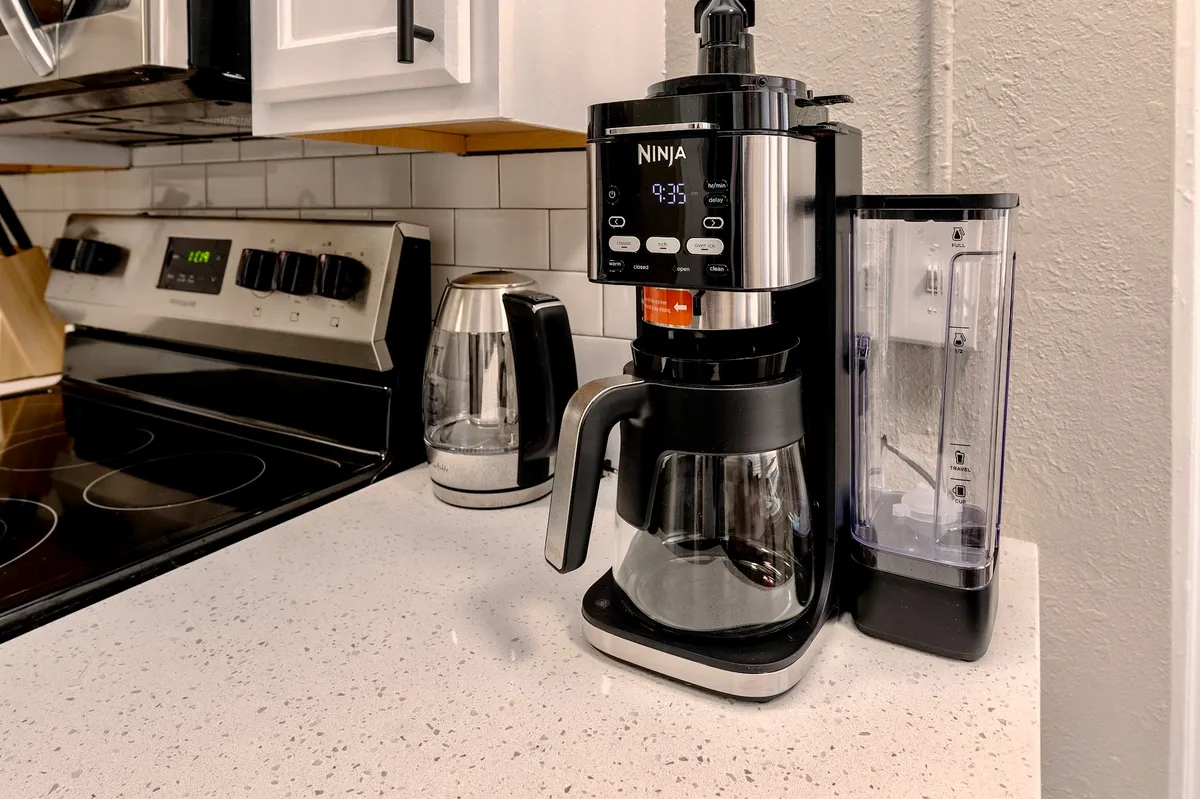 A detailed look at kitchen essentials including a Ninja coffee maker, stainless steel kettle, and stove, all set against a clean, functional backdrop. The space exudes a cozy, practical vibe ideal for casual meal prep or brewing morning coffee.