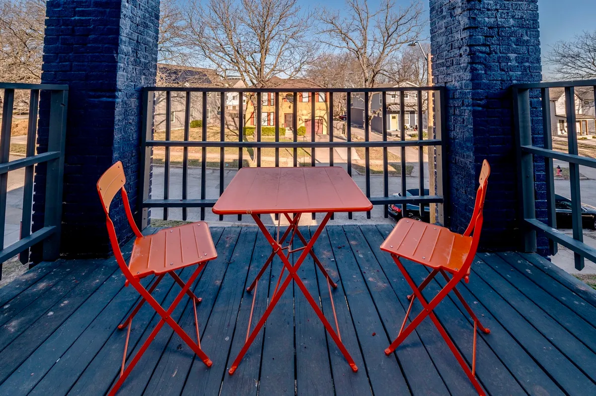 A cheerful exterior patio area with bright orange tables and chairs overlooks a quiet street, suggesting a welcoming community environment. The setting is bathed in warm sunlight, offering a glimpse of the surrounding neighborhood and hinting at the apartment’s close proximity to local amenities.