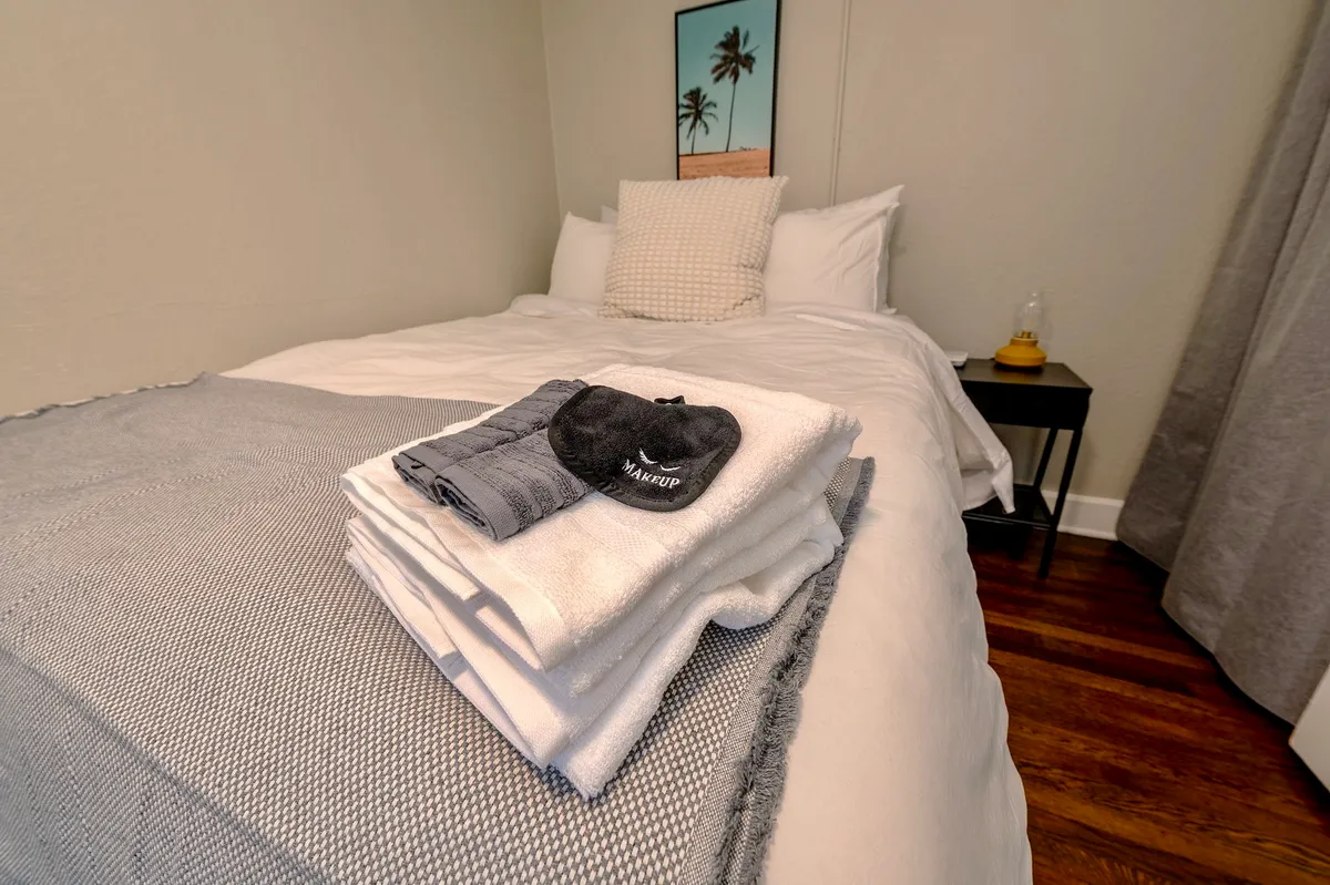 A cozy bedroom featuring a neatly made bed with soft linens, a decorative pillow, a stack of plush towels at the foot, and a nightstand. The overall mood is tranquil and inviting, emphasizing comfort and relaxation.