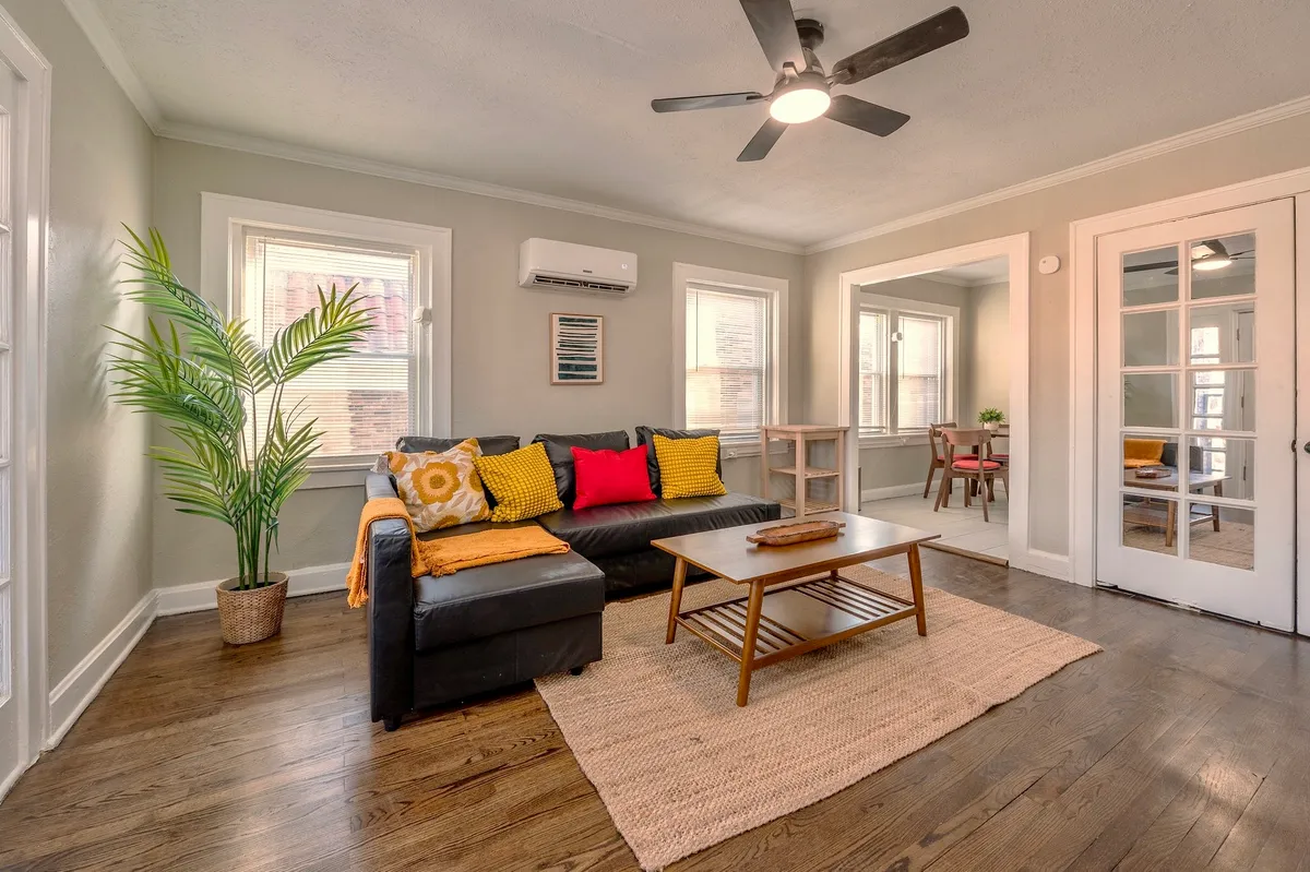 This scene displays a black sectional sofa with colorful cushions, a wooden coffee table, and an indoor plant, set beside glass doors that draw in natural light and open to another room. The arrangement is both welcoming and functional, ideal for socializing, relaxing, or reading in a light-filled setting.