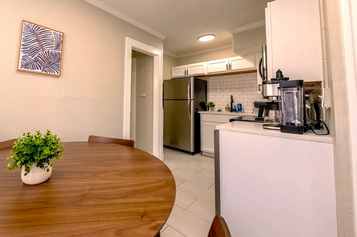An inviting dining nook with a round wooden table set near a stainless steel refrigerator and white cabinetry. Framed artwork adds a touch of style, while the arrangement feels intimate and suitable for casual meals or morning coffee.